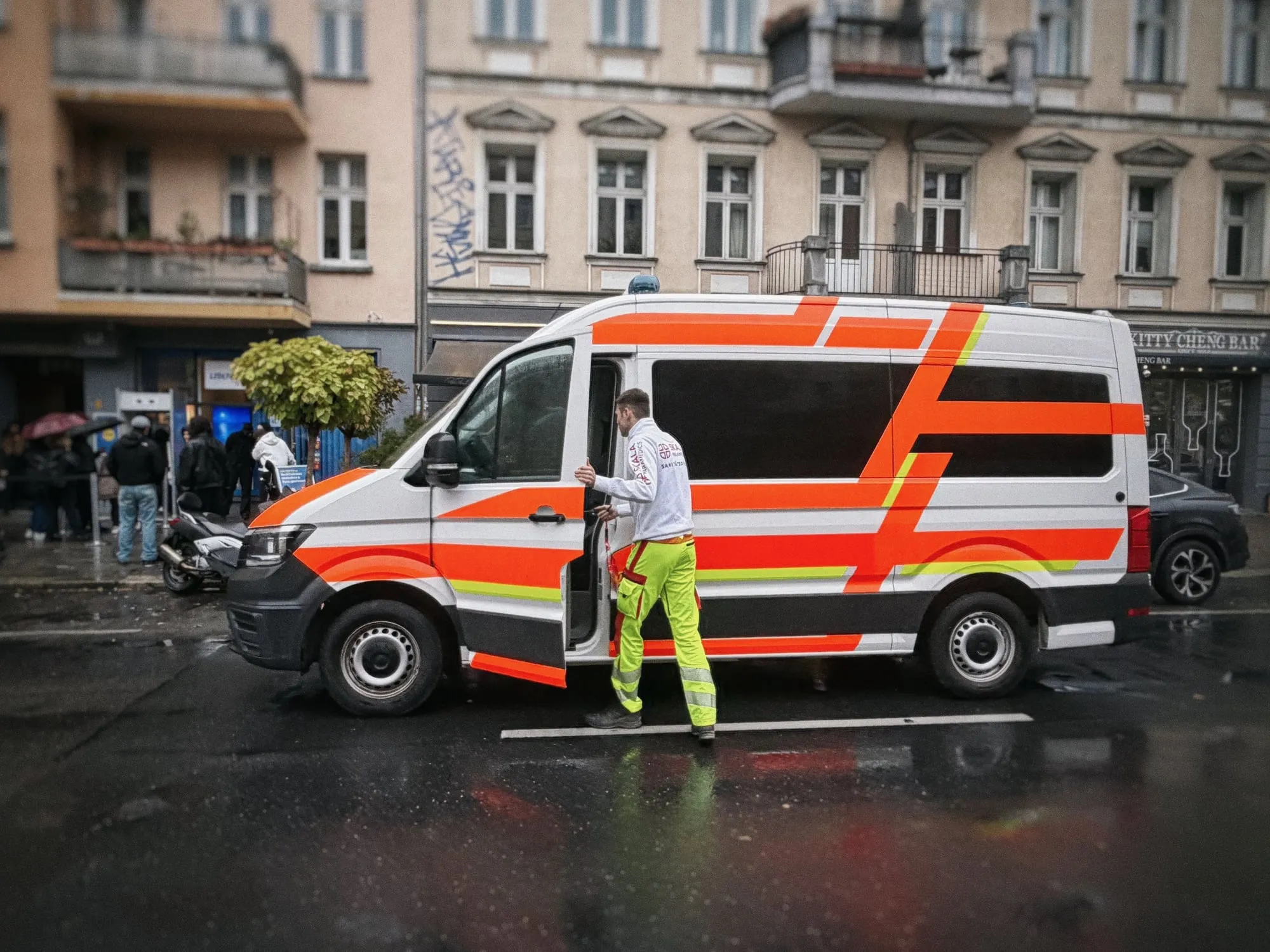 Sanitäter steigt bei Regen in den Krankentransportwagen von SKALA Paramedics, im Hintergrund die Warteschlange vor „Lidlton Reisen“