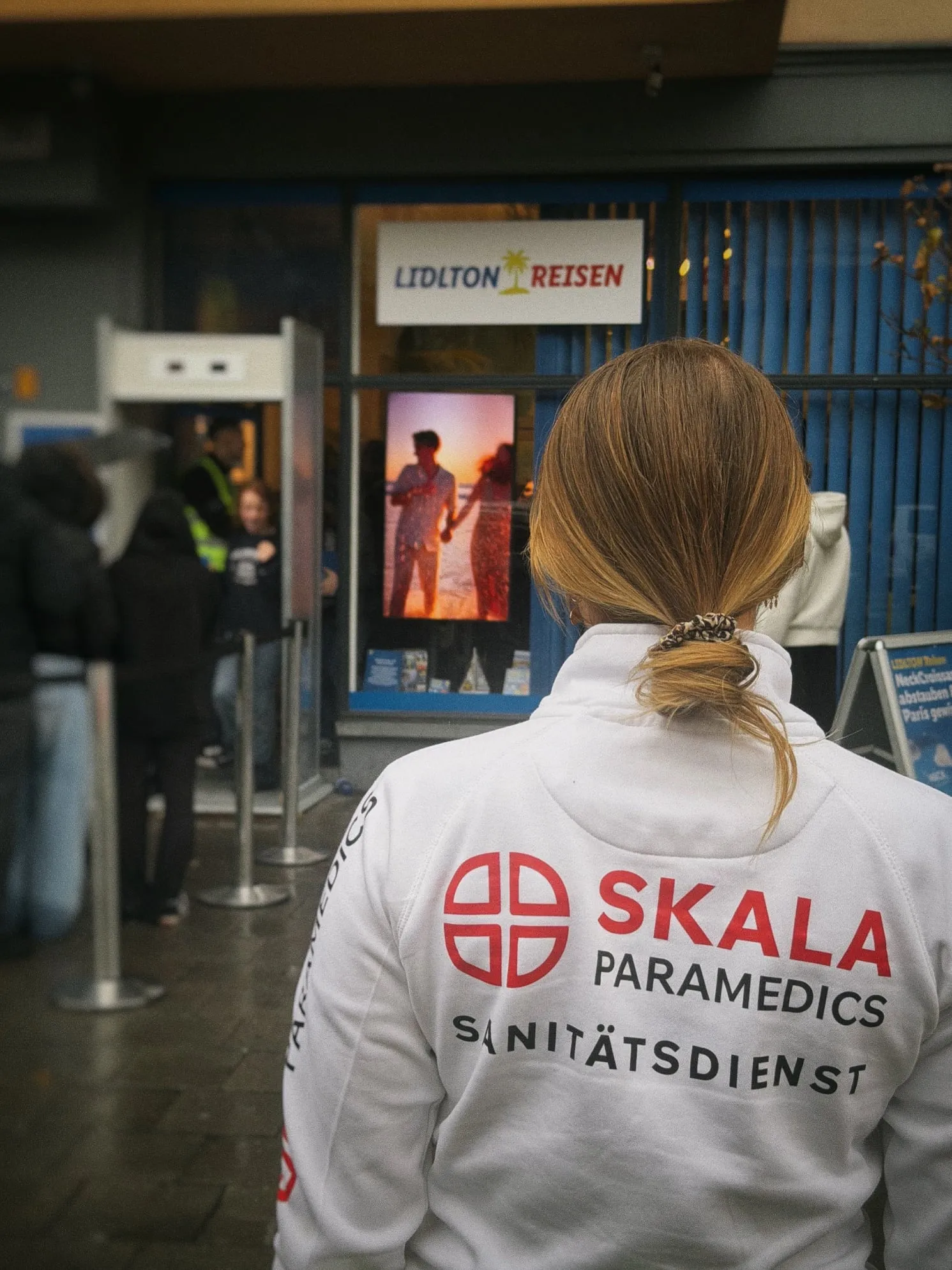 Sanitäterin von SKALA Paramedics von hinten vor dem Pop-up-Store „Lidlton Reisen“ mit Warteschlange und Einlasskontrolle
