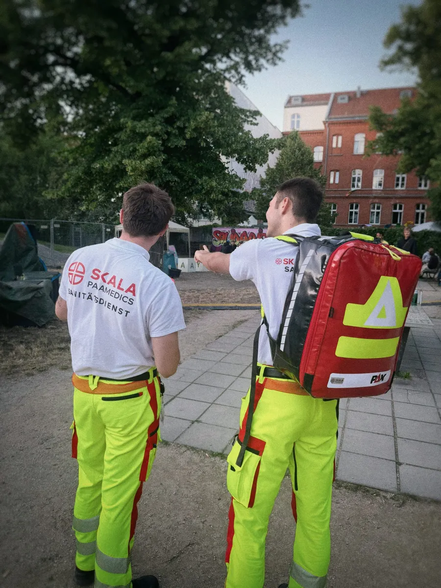 Zwei Sanitäter mit rotem Notfallrucksack bei einem Outdoor-Event, von hinten